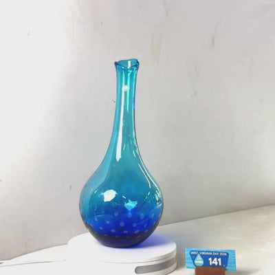 A translucent blue glass vase with a flared rim and dotted pattern near the base, displayed on a white circular stand against a plain light gray wall.