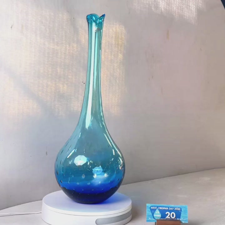 A translucent blue glass vase with a flared neck and dotted pattern, displayed on a white circular base against a light gray wall.