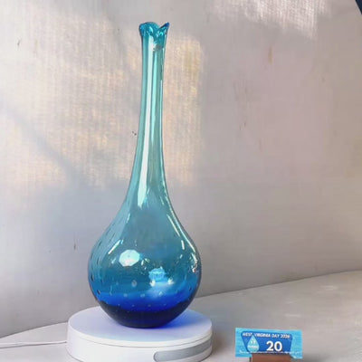 A translucent blue glass vase with a flared neck and dotted pattern, displayed on a white circular base against a light gray wall.