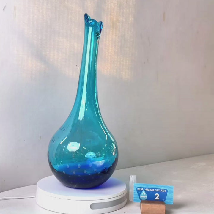 A translucent blue glass vase with a flared neck and dotted pattern, displayed on a white circular base against a light gray wall.