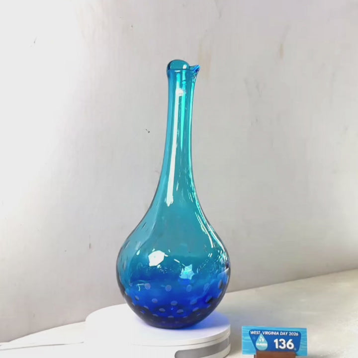 A translucent blue glass vase with a flared rim and dotted pattern near the base, displayed on a white circular stand against a plain light gray wall.