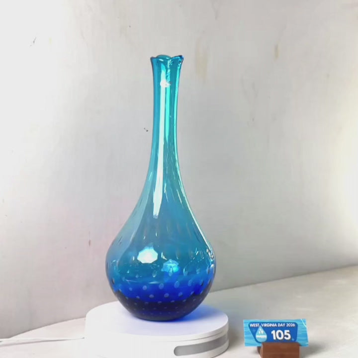 A translucent blue glass vase with a flared rim and dotted pattern near the base, displayed on a white circular stand against a plain light gray wall.