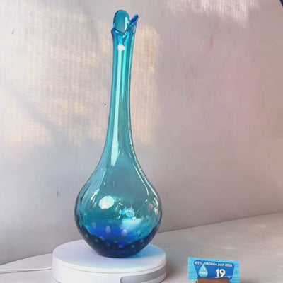 A translucent blue glass vase with a flared neck and dotted pattern, displayed on a white circular base against a light gray wall.