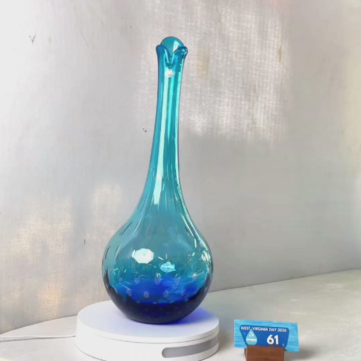 A translucent blue glass vase with a flared neck and dotted pattern, displayed on a white circular base against a light gray wall.