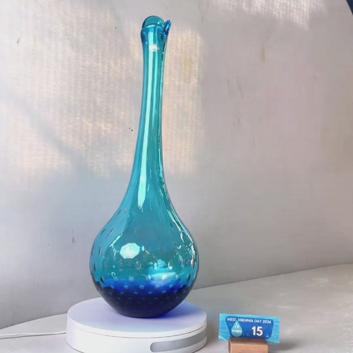 A translucent blue glass vase with a flared neck and dotted pattern, displayed on a white circular base against a light gray wall.