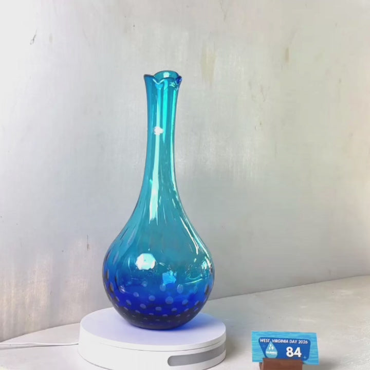 A translucent blue glass vase with a flared rim and subtle white speckled pattern near the base, displayed on a white round scale with a small price tag in front, against a plain light gray wall.