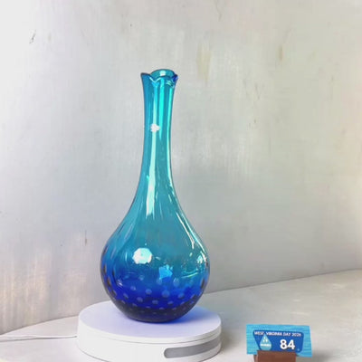 A translucent blue glass vase with a flared rim and subtle white speckled pattern near the base, displayed on a white round scale with a small price tag in front, against a plain light gray wall.