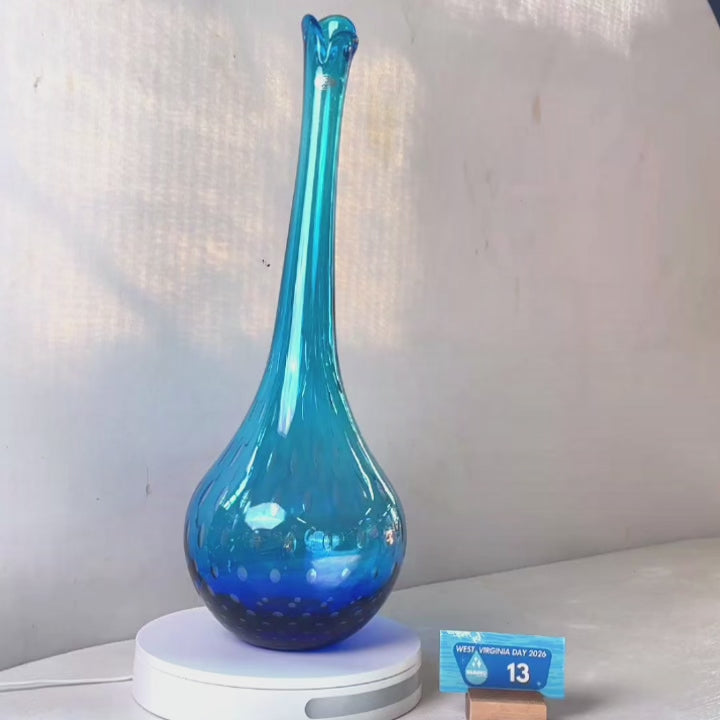 A translucent blue glass vase with a flared neck and dotted pattern, displayed on a white circular base against a light gray wall.