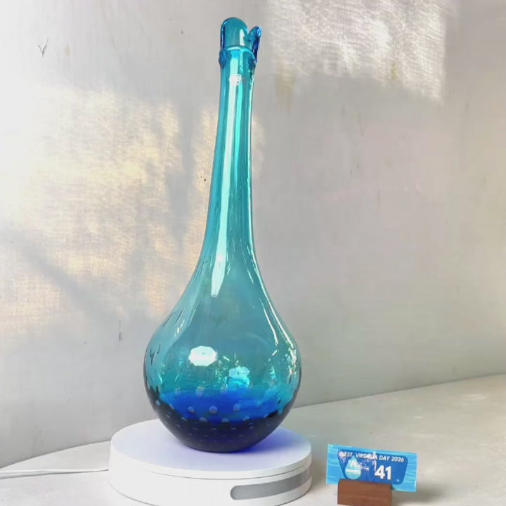A translucent blue glass vase with a flared neck and dotted pattern, displayed on a white circular base against a light gray wall.