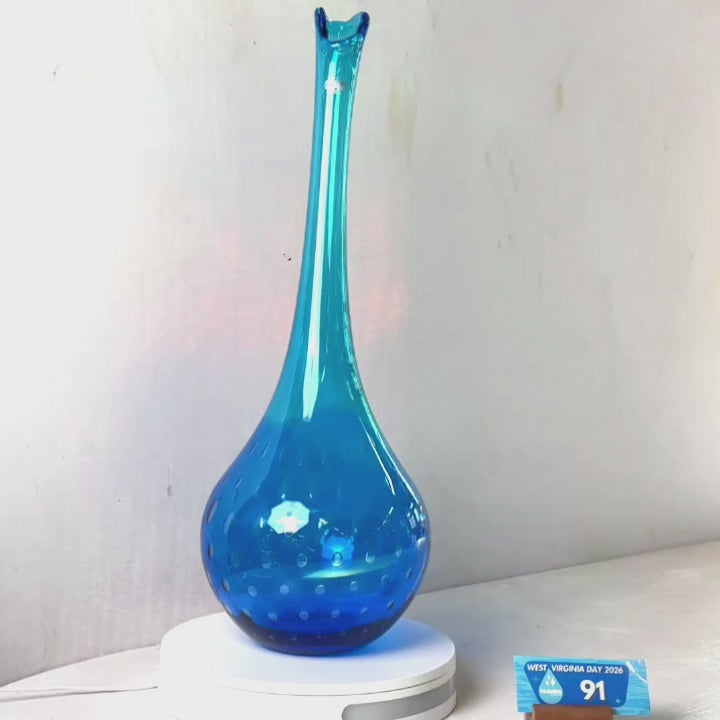 A translucent blue glass vase with a flared rim and subtle white speckled pattern near the base, displayed on a white round scale with a small price tag in front, against a plain light gray wall.