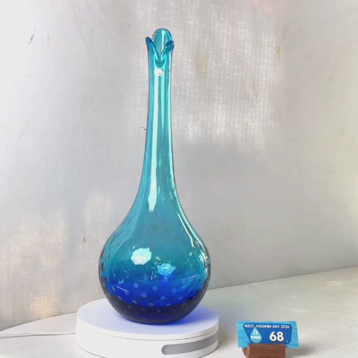 A translucent blue glass vase with a flared neck and dotted pattern, displayed on a white circular base against a light gray wall.