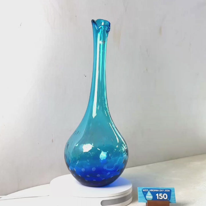 A translucent blue glass vase with a flared rim and dotted pattern near the base, displayed on a white circular stand against a plain light gray wall.