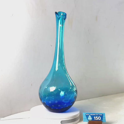 A translucent blue glass vase with a flared rim and dotted pattern near the base, displayed on a white circular stand against a plain light gray wall.