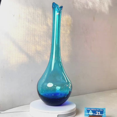 A translucent blue glass vase with a flared neck and dotted pattern, displayed on a white circular base against a light gray wall.
