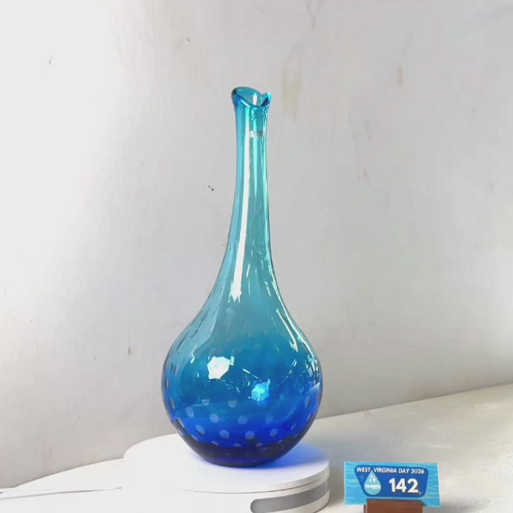 A translucent blue glass vase with a flared rim and dotted pattern near the base, displayed on a white circular stand against a plain light gray wall.
