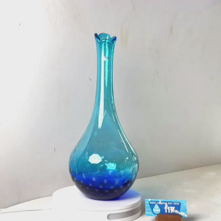 A translucent blue glass vase with a flared neck and dotted pattern near the base, displayed on a white circular light box. A small wrapped item and a brown block are placed beside it on a white surface against a plain light gray wall.