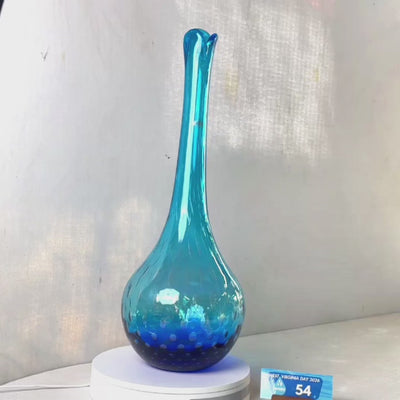 A translucent blue glass vase with a flared neck and dotted pattern, displayed on a white circular base against a light gray wall.