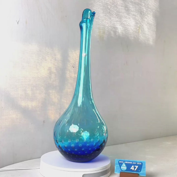 A translucent blue glass vase with a flared neck and dotted pattern, displayed on a white circular base against a light gray wall.