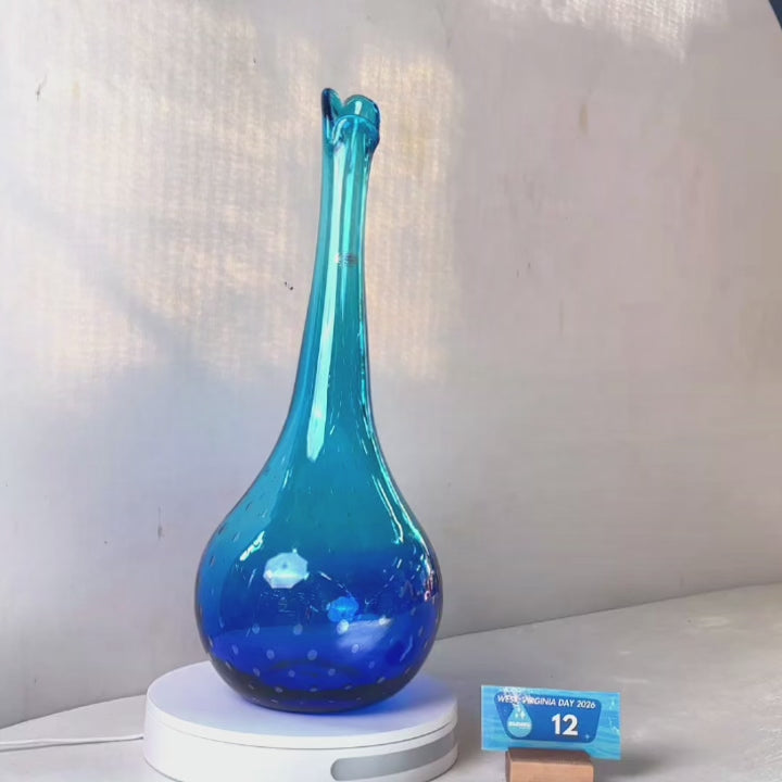 A translucent blue glass vase with a flared neck and dotted pattern, displayed on a white circular base against a light gray wall.