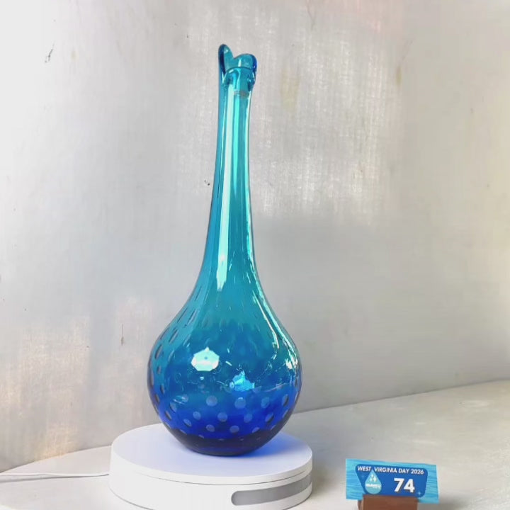 A translucent blue glass vase with a flared rim and subtle white speckled pattern near the base, displayed on a white round scale with a small price tag in front, against a plain light gray wall.