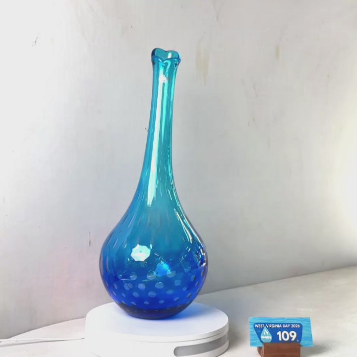 A translucent blue glass vase with a flared rim and dotted pattern near the base, displayed on a white circular stand against a plain light gray wall.