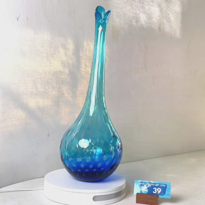 A translucent blue glass vase with a flared neck and dotted pattern, displayed on a white circular base against a light gray wall.