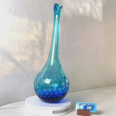 A translucent blue glass vase with a flared neck and dotted pattern, displayed on a white circular base against a light gray wall.