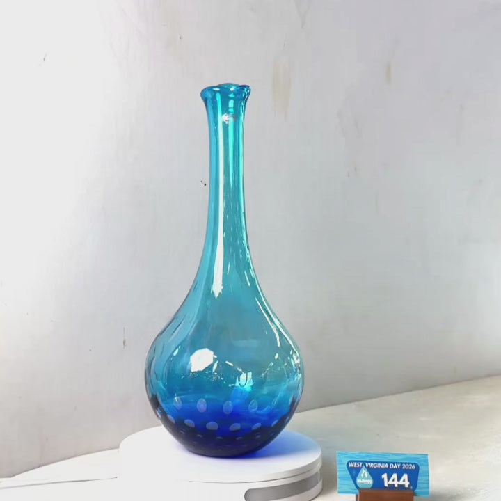 A translucent blue glass vase with a flared rim and dotted pattern near the base, displayed on a white circular stand against a plain light gray wall.