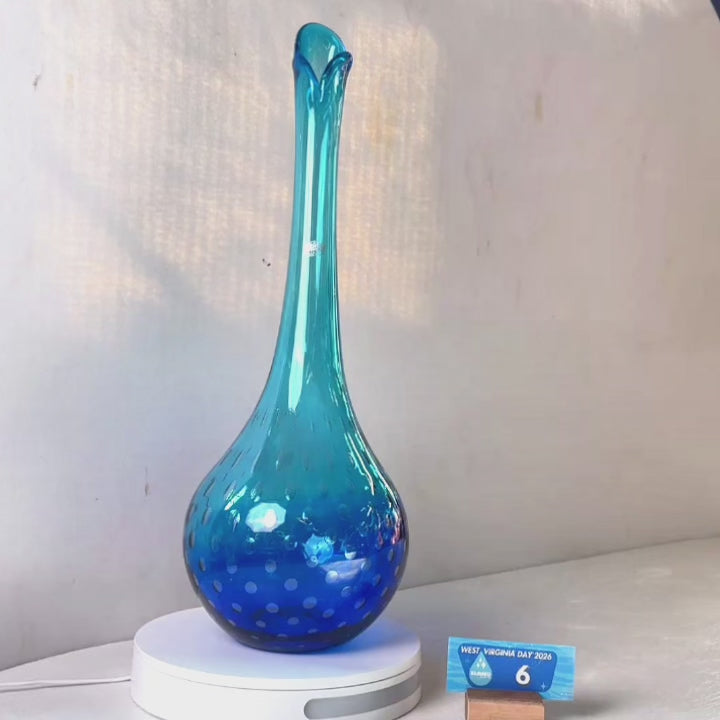A translucent blue glass vase with a flared neck and dotted pattern, displayed on a white circular base against a light gray wall.