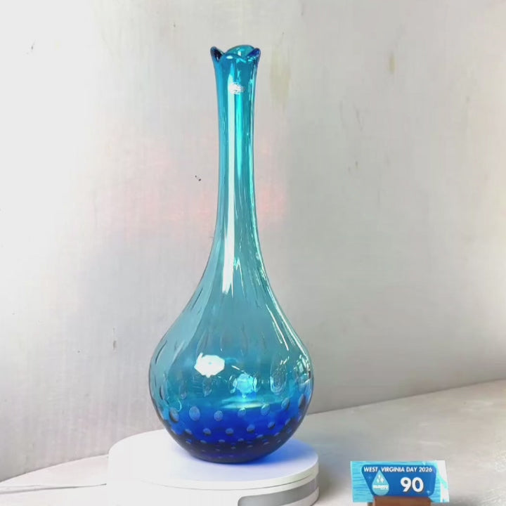 A translucent blue glass vase with a flared rim and subtle white speckled pattern near the base, displayed on a white round scale with a small price tag in front, against a plain light gray wall.