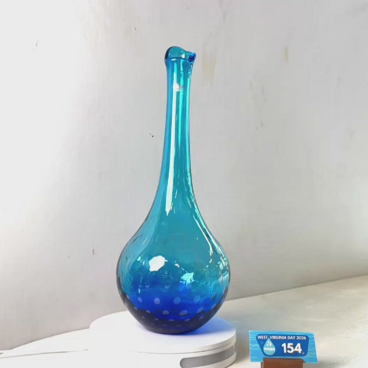 A translucent blue glass vase with a flared rim and dotted pattern near the base, displayed on a white circular stand against a plain light gray wall.