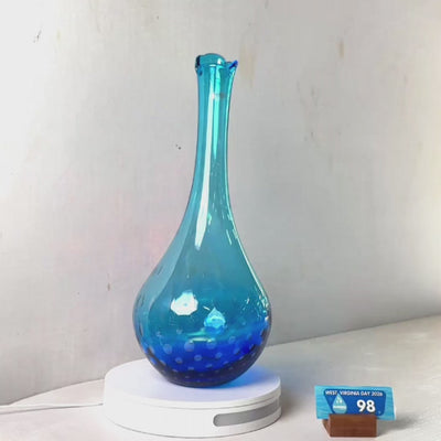 A tall, slender cobalt blue glass vase with a flared neck and small white polka dots, displayed on a white circular light box against a plain light gray wall.
