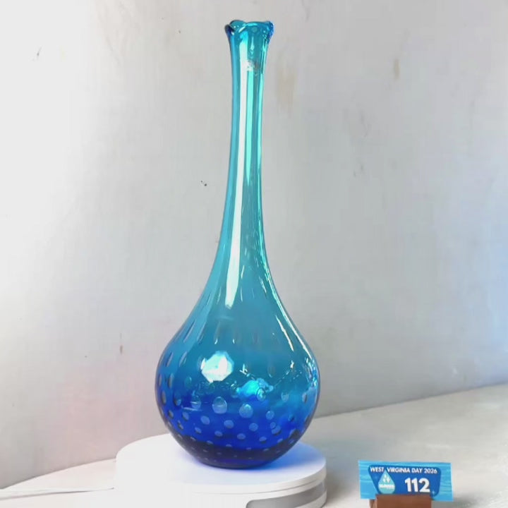 A translucent blue glass vase with a flared rim and dotted pattern near the base, displayed on a white circular stand against a plain light gray wall.