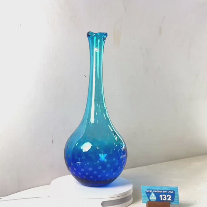 A translucent blue glass vase with a flared rim and dotted pattern near the base, displayed on a white circular stand against a plain light gray wall.