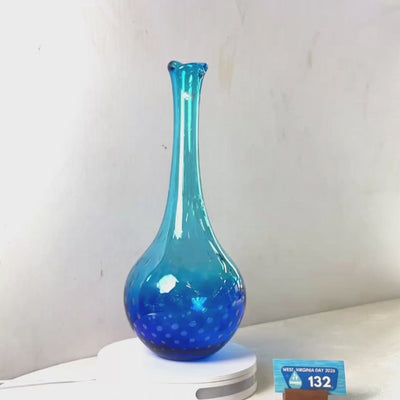 A translucent blue glass vase with a flared rim and dotted pattern near the base, displayed on a white circular stand against a plain light gray wall.