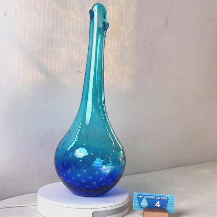 A translucent blue glass vase with a flared neck and dotted pattern, displayed on a white circular base against a light gray wall.