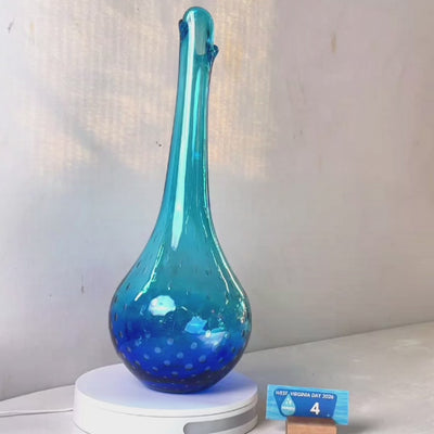 A translucent blue glass vase with a flared neck and dotted pattern, displayed on a white circular base against a light gray wall.