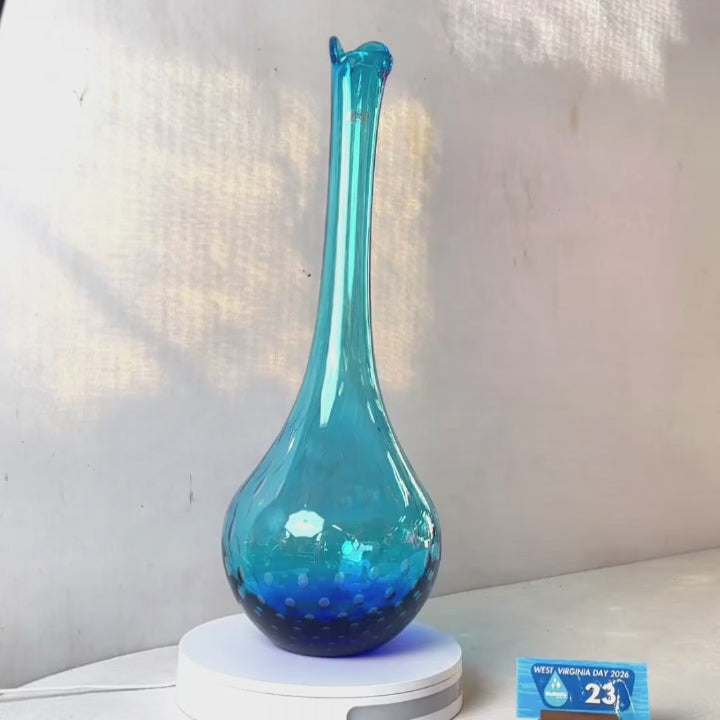 A translucent blue glass vase with a flared neck and dotted pattern, displayed on a white circular base against a light gray wall.