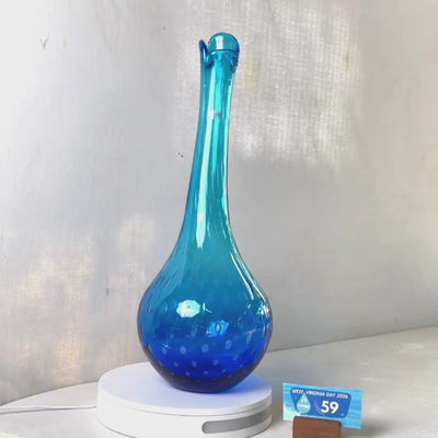 A translucent blue glass vase with a flared neck and dotted pattern, displayed on a white circular base against a light gray wall.
