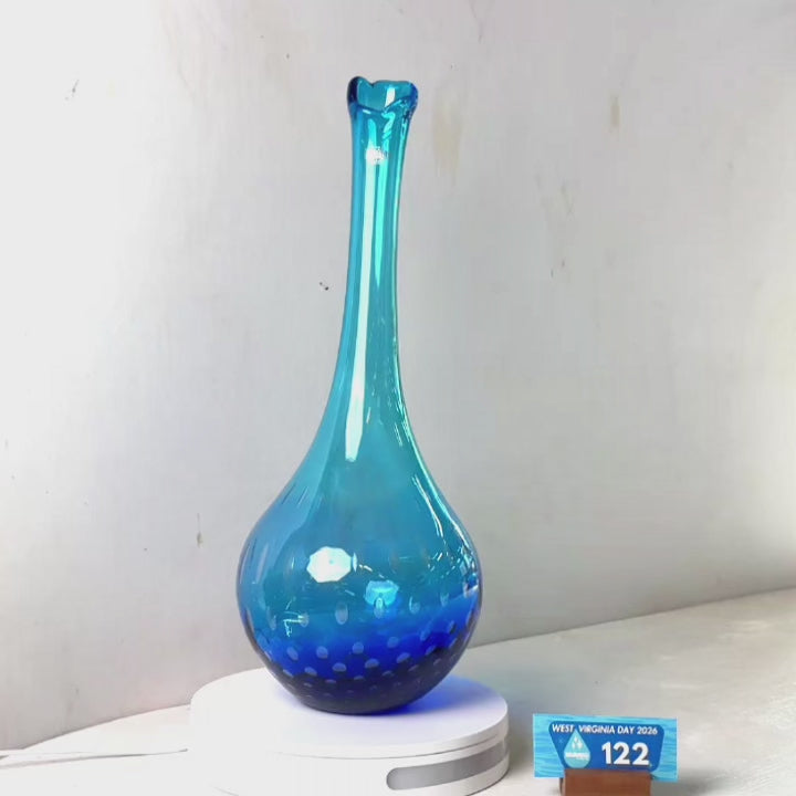 A translucent blue glass vase with a flared neck and dotted pattern near the base, displayed on a white circular light box. A small wrapped item and a brown block are placed beside it on a white surface against a plain light gray wall.