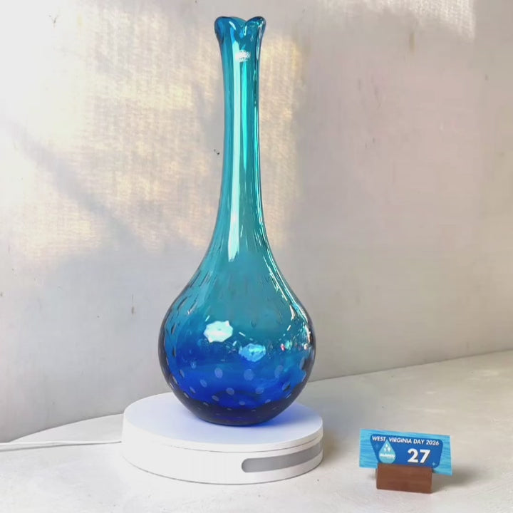 A translucent blue glass vase with a flared neck and dotted pattern, displayed on a white circular base against a light gray wall.