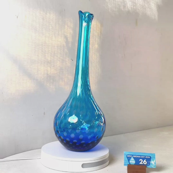 A translucent blue glass vase with a flared neck and dotted pattern, displayed on a white circular base against a light gray wall.