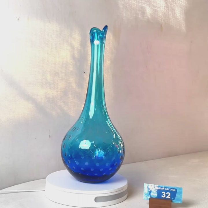 A translucent blue glass vase with a flared neck and dotted pattern, displayed on a white circular base against a light gray wall.