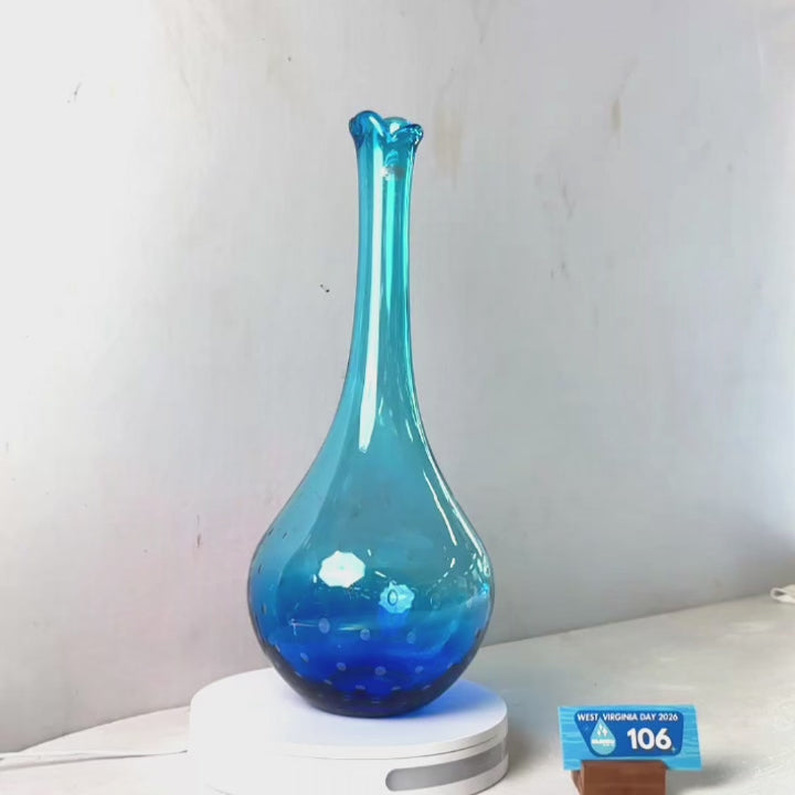 A translucent blue glass vase with a flared rim and dotted pattern near the base, displayed on a white circular stand against a plain light gray wall.