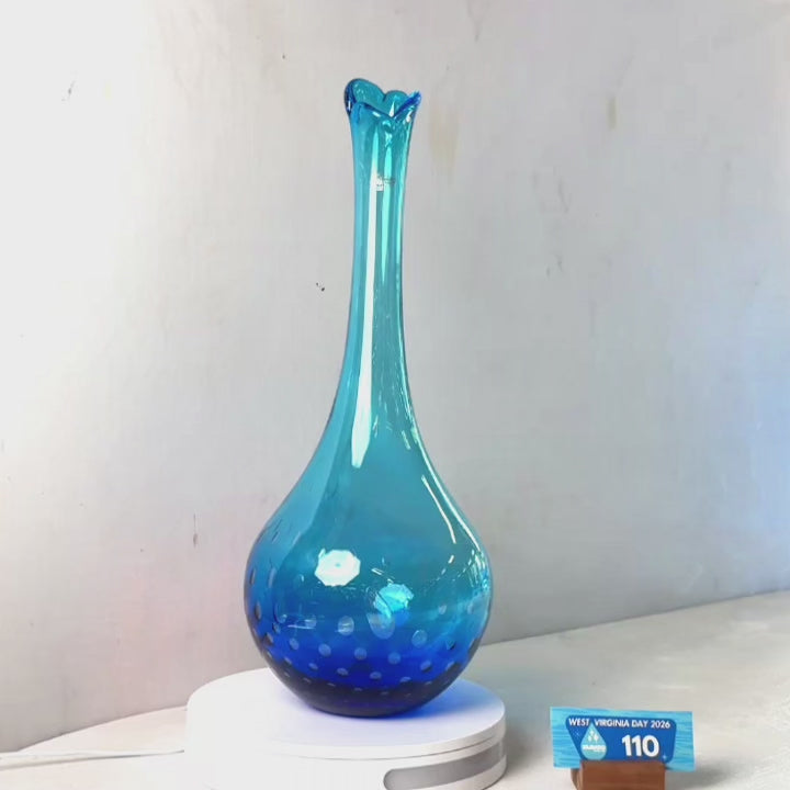 A translucent blue glass vase with a flared rim and dotted pattern near the base, displayed on a white circular stand against a plain light gray wall.