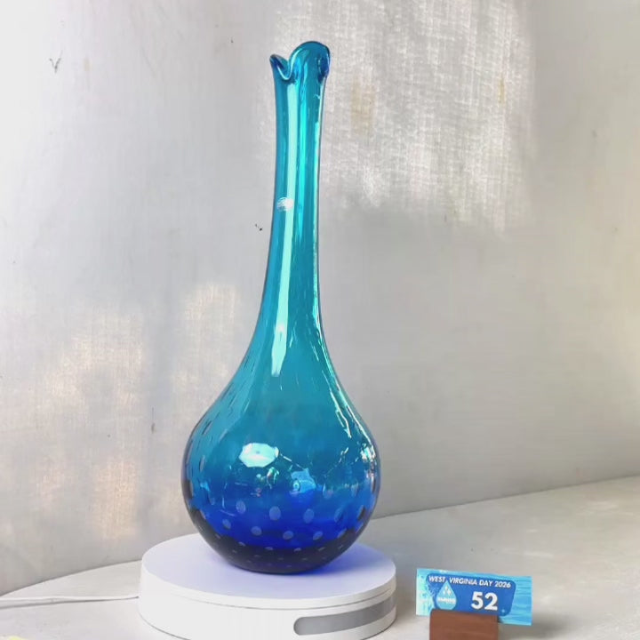 A translucent blue glass vase with a flared neck and dotted pattern, displayed on a white circular base against a light gray wall.