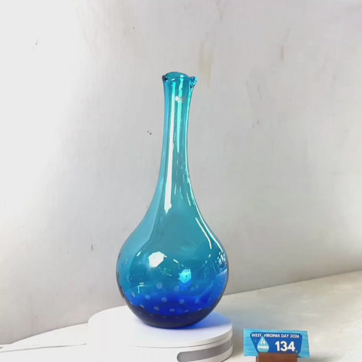 A translucent blue glass vase with a flared rim and dotted pattern near the base, displayed on a white circular stand against a plain light gray wall.