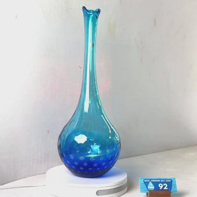 A translucent blue glass vase with a flared rim and subtle white speckled pattern near the base, displayed on a white round scale with a small price tag in front, against a plain light gray wall.
