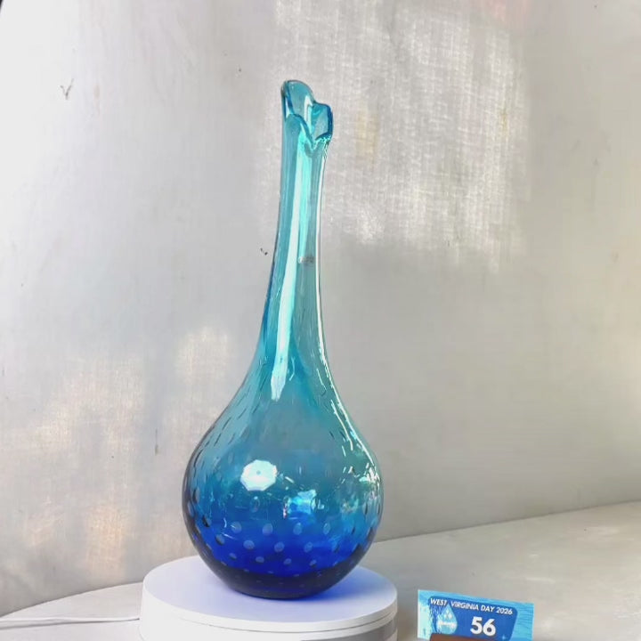 A translucent blue glass vase with a flared neck and dotted pattern, displayed on a white circular base against a light gray wall.
