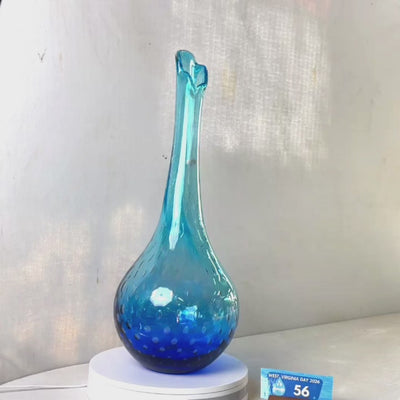 A translucent blue glass vase with a flared neck and dotted pattern, displayed on a white circular base against a light gray wall.